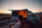 A person sits inside a lit van in a desert at sunset. An open sleeping bag is outside, creating a cozy, adventurous atmosphere.