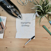 Crumpled paper labeled Marketing Strategy on wooden table with iron, books, plant, tape, markers, and blue ribbons, suggesting a work setup.