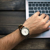 Person wearing a brown watch types on a laptop keyboard. The setting is a wooden desk. The mood is focused.