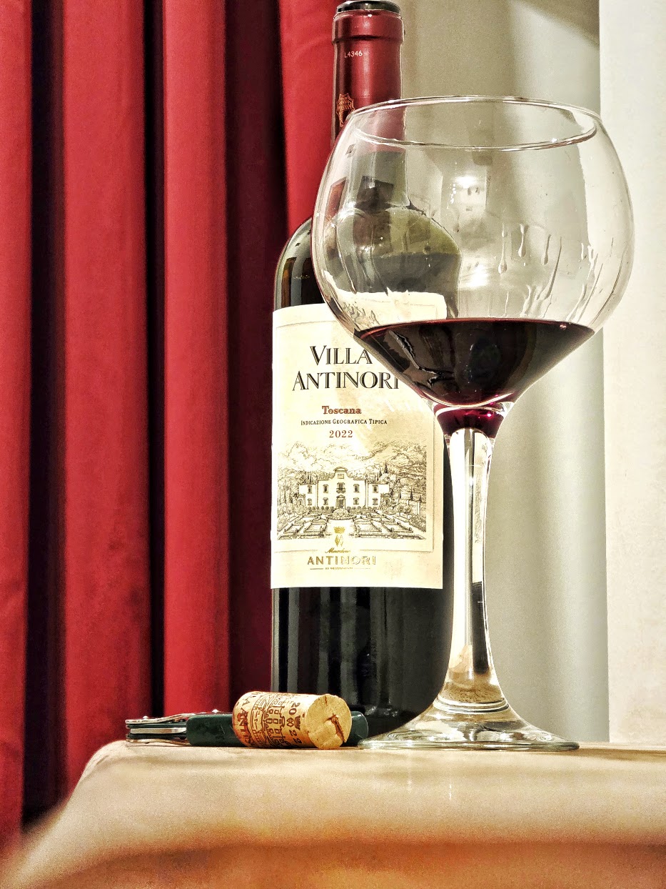 Bouteille de Villa Antinori Rosso débouchonnée à La Corte Segreta, avec un verre de vin rouge, son bouchon et un tire-bouchon devant un rideau rouge.