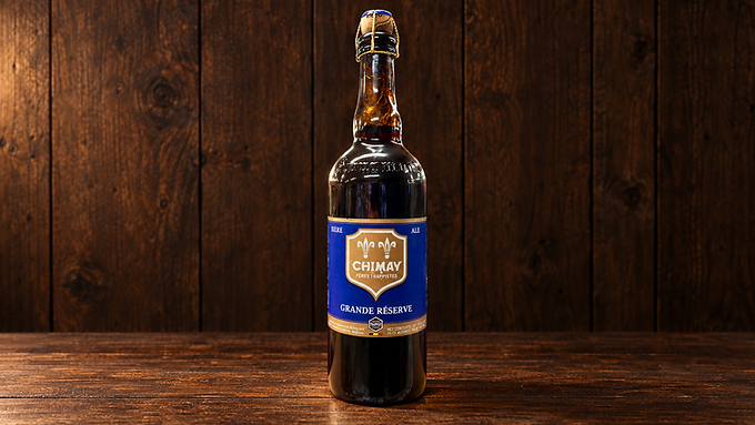 Bouteille de Chimay Bleue Grande Réserve 75 cl, à étiquette bleue et dorée, sur une table en bois devant un mur en bois.