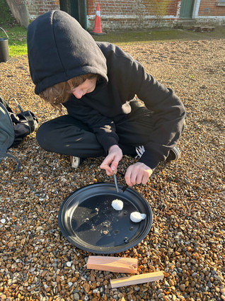 A Scout sits on gravel with a metal plate in front of him. On top are two cotton wool balls and he is using a ferro rod to try and ignite them. In front of the metal plate are two pieces of kindling wood