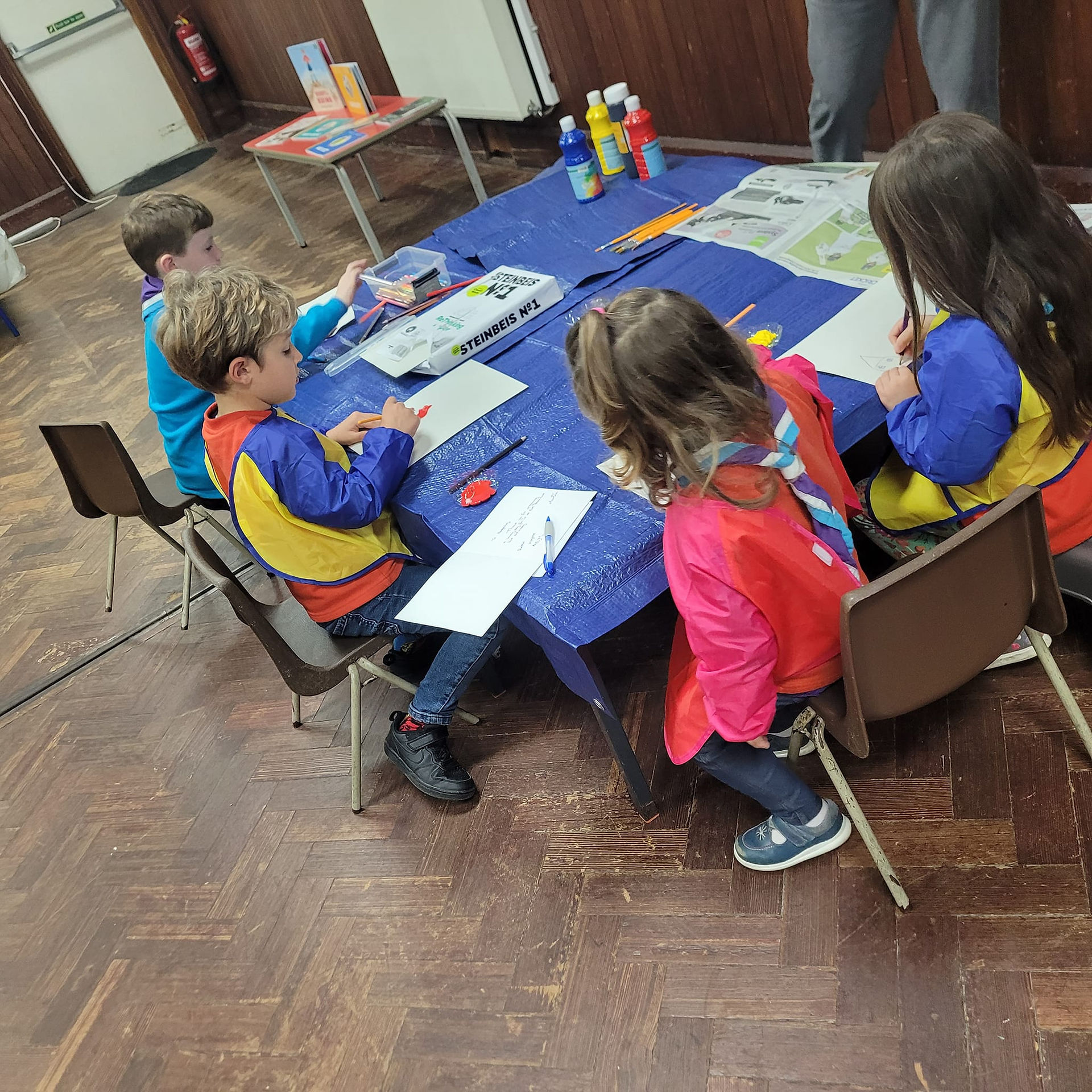 Squirrel and Beaver Scouts sit around a table wearing aprons. There are bottles of paint on the table with their brushes and paper on a plastic table cloth
