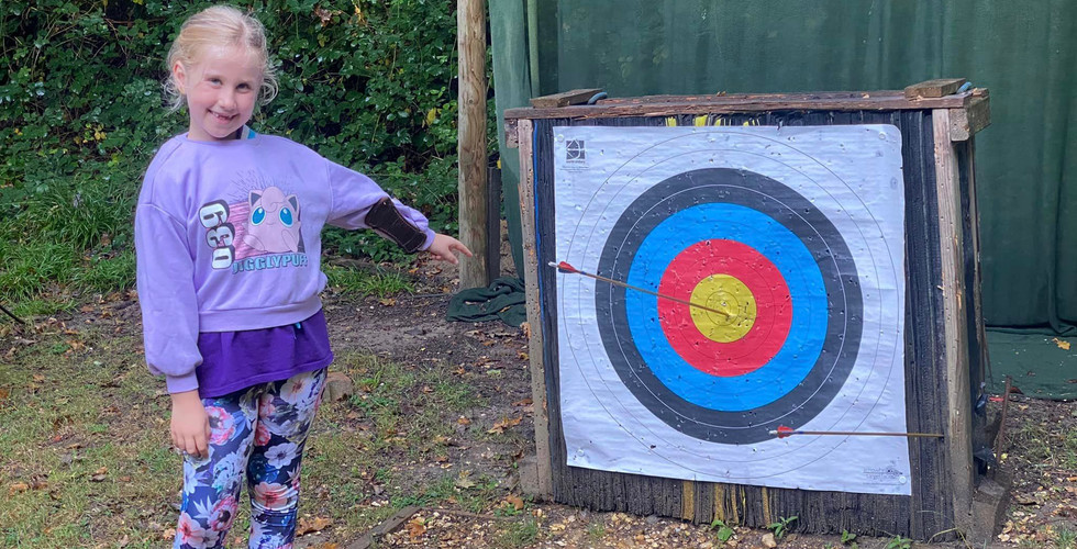 A Beaver Scout points at an archery target. One arrow is off the target on the wooden frame and the other is in the centre circle of the target