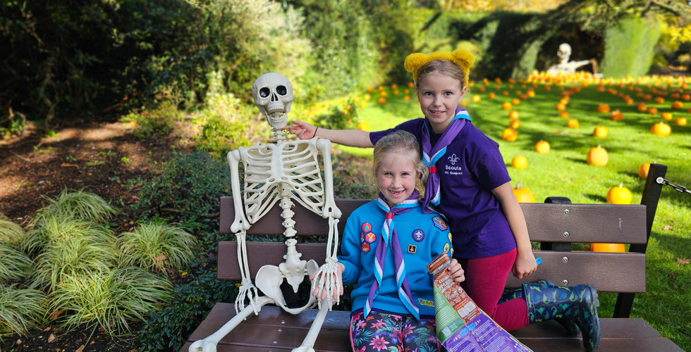 Two Beavers sit on a bench next to a plastic skeleton. There are pumpkins in the field behind them and one of the Beavers is holding an unfolded map
