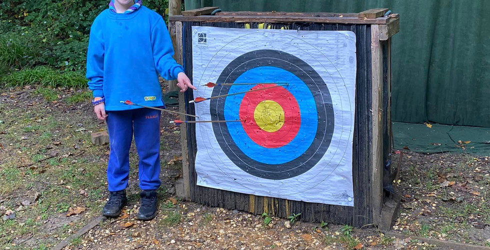 A Beaver Scout points to an archery target. There is one arrow outside of the target but on the paper, two in the circle second from the outside of the target which are right next to each other and a fourth one in the second from the centre circle of the target