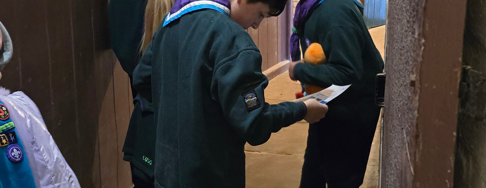 Three Cub Scouts stand in a narrow corridor with an adult volunteer. A Beaver Scout is partially visible on the left of the image. The walls are lined with wood panelled arch shaped walls between brick arches. Each wooden panel has a doorway. The floor is stone