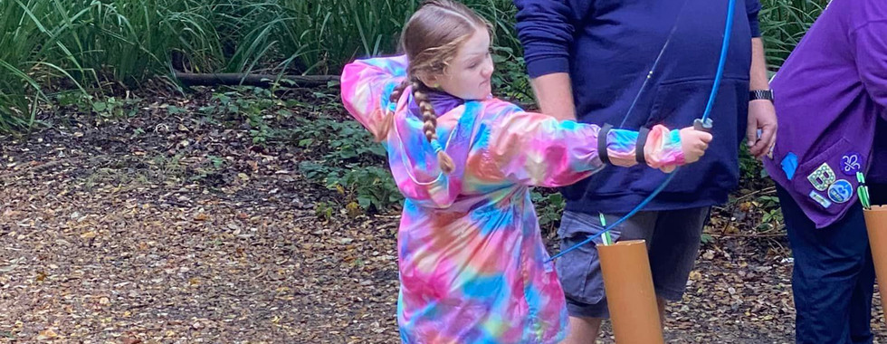 A Beaver Scout holds an archery bow with an adult stood next to her supervising. Arrows are in a drainpipe in front of her sunk into the floor