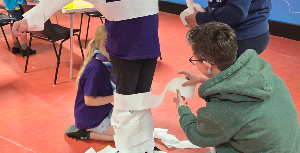 An adult helps a Cub and a Scout wrap another Cub in toilet paper as part of a game