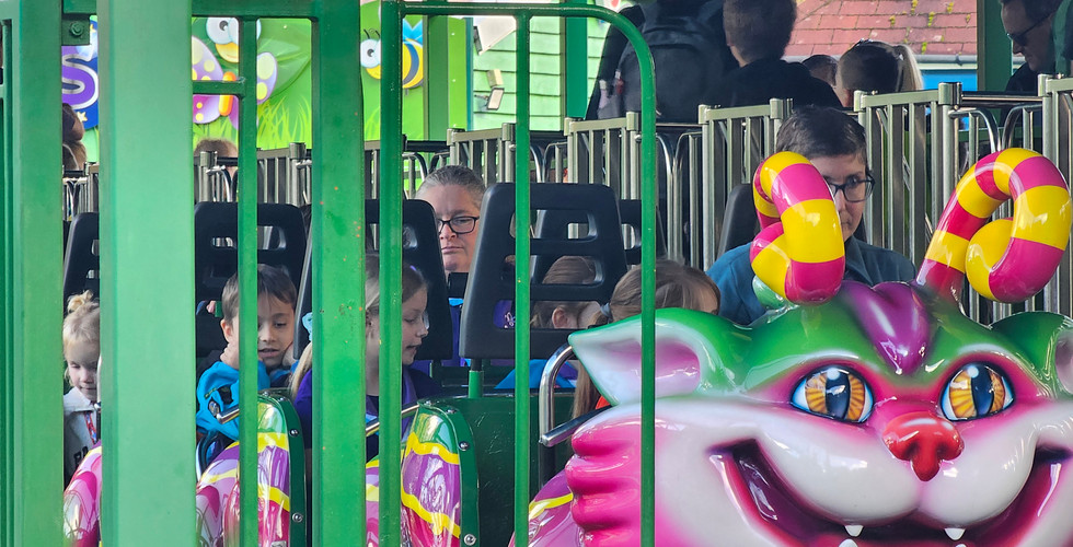 A roller coaster that looks like a cat with caterpillar antennae. From the front carriage backwards the image is obscured by green fencing but some Beaver Scouts are visible in the seats behind the bars