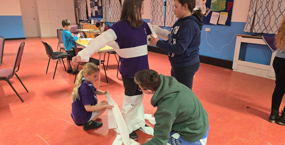 An adult helps a Cub and a Scout wrap another Cub in toilet paper as part of a game
