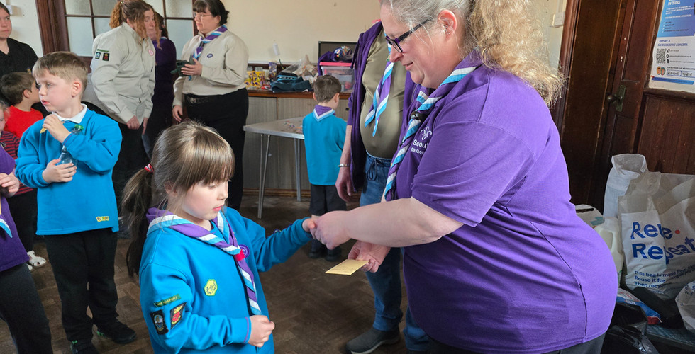 A Beaver Scout shakes left hands with an adult who hands her an envelope containing her badges