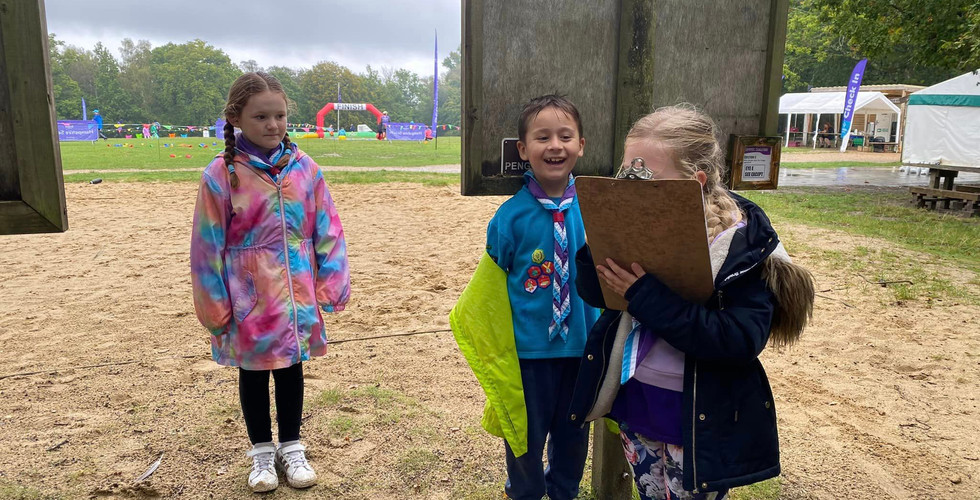 Three Beaver Scouts stand in a sandy area behind a wooden sign post, one holds a clipboard