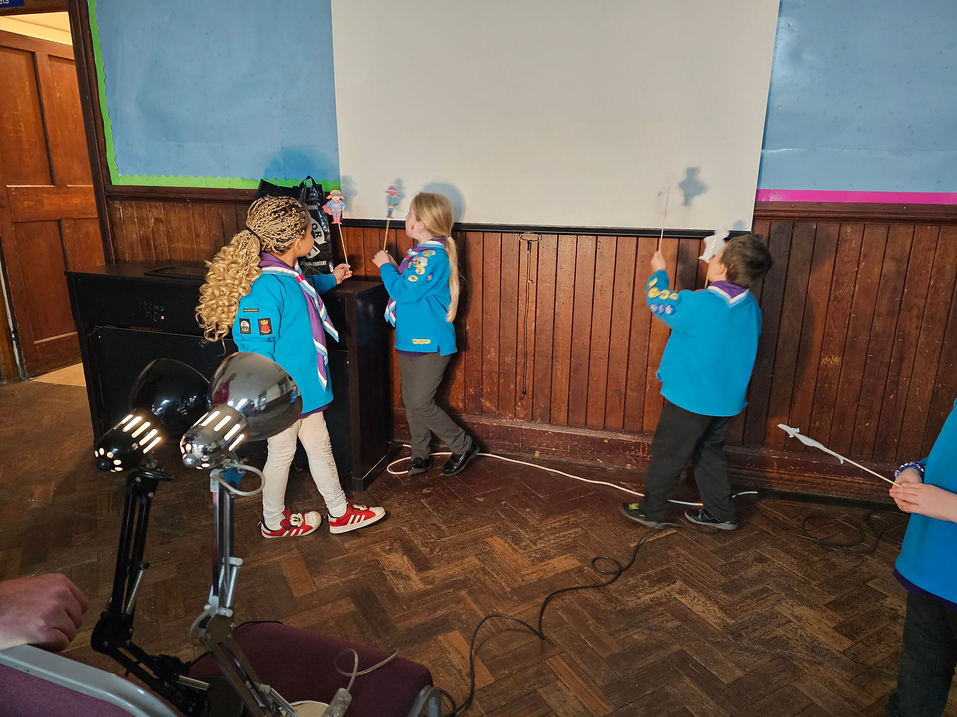 Some Beaver Scouts stand between a projection screen and some lamps to play with their shadow puppet superheroes