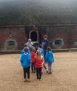 Squirrel and Beaver Scouts stand in front of their group's volunteer and a Young Leader. Behind them are iron railings and a Victorian fort. They are standing on gravel