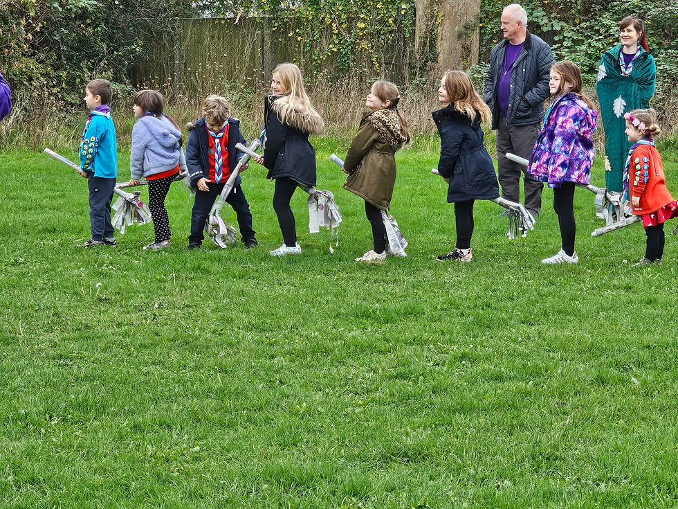A group of Beaver and Squirrel Scouts sit on the newspaper broomsticks they made in a field with some of their leaders nearby
