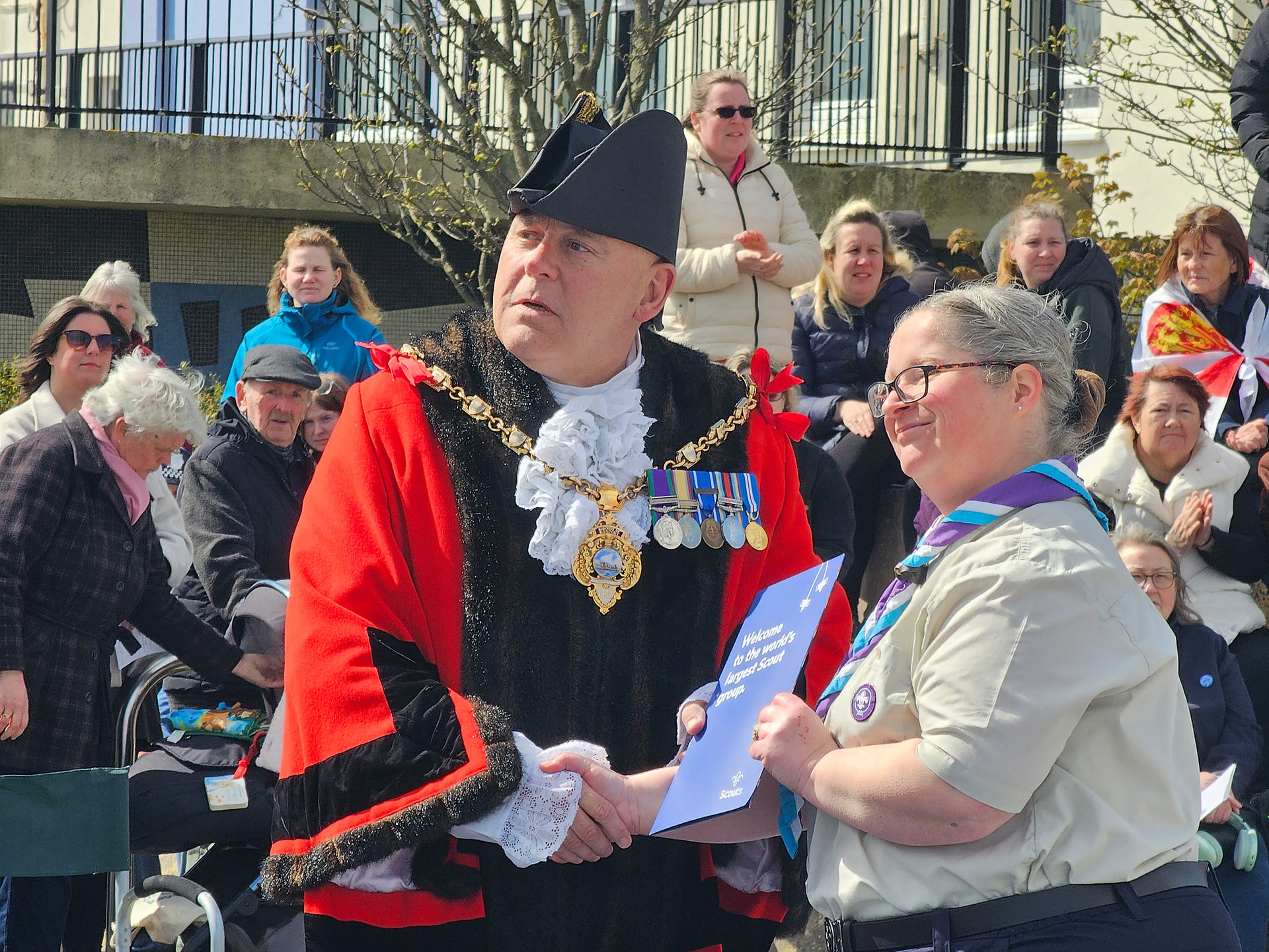 A Scout leader poses with the Mayor holding a certificate between them, behind them are spectators