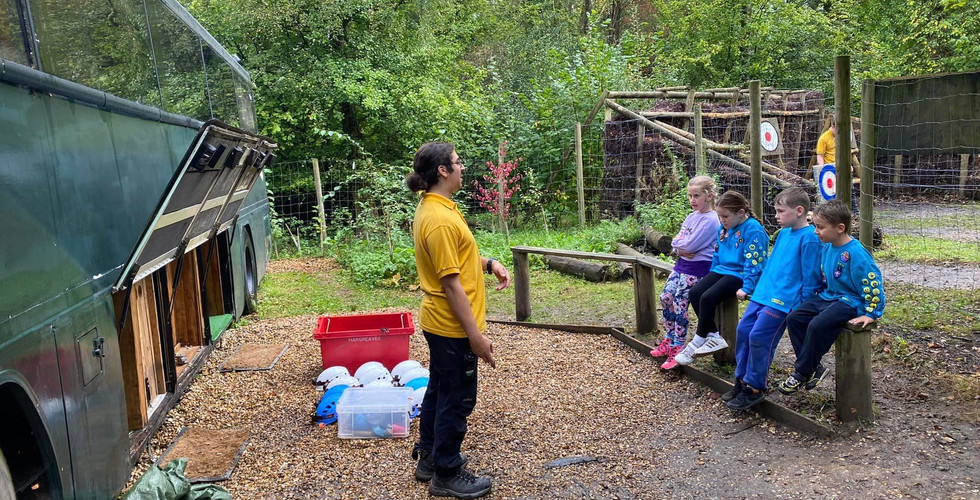Four Beaver Scouts sit on a wooden bench listening to a leader who stands in front of them, behind the leader is a bus with its side open. On the floor are hard hats, boxes and tarpaulins