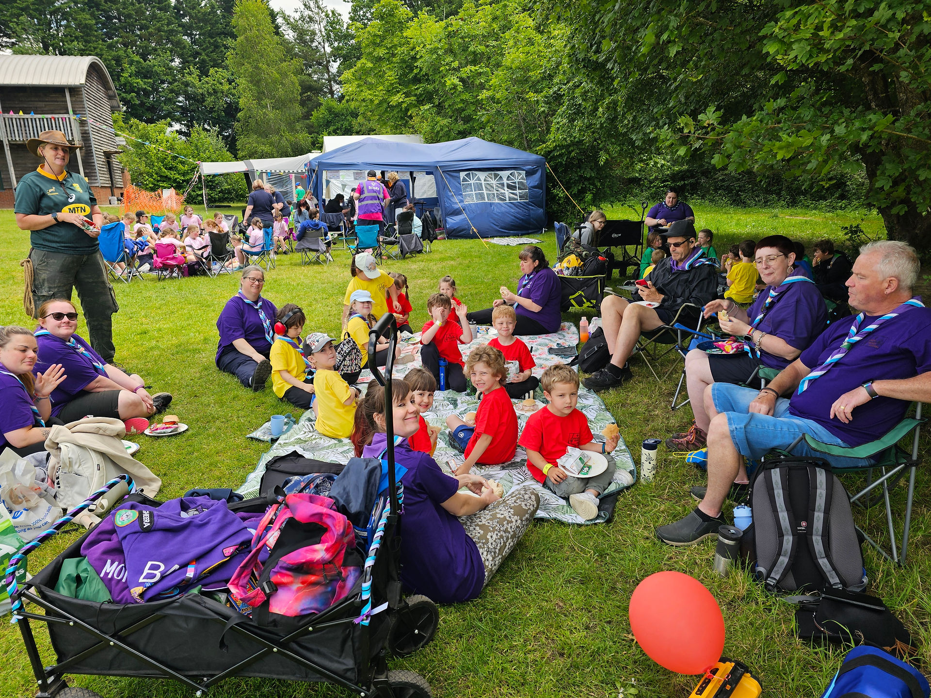 Squirrel and Beaver Scouts sit with their leaders under a tree. Most of them are sat on picnic blankets but some leaders are sat on camp chairs. There's a camp trolley in front of them all with their bags in