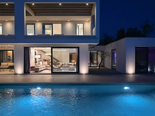 Modern luxury home at night with glowing pool, open patio, and lit interior rooms under a deep blue sky.