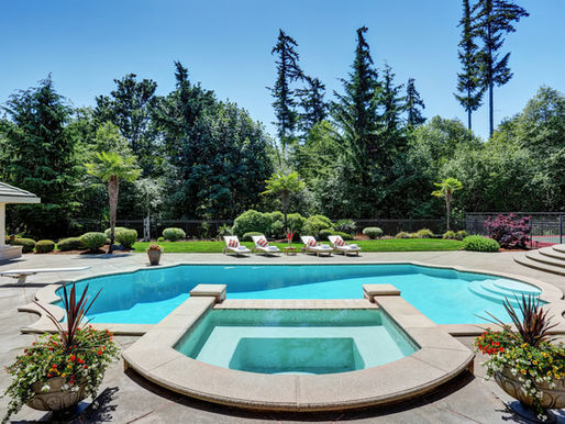 Luxurious backyard pool and hot tub with lounge chairs, flowers, and tall trees under a clear blue sky