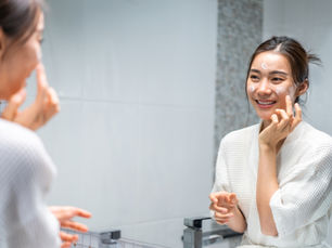 A woman in a white robe applies skincare cream, smiling in front of a bathroom mirror. The setting is bright with a tiled background.