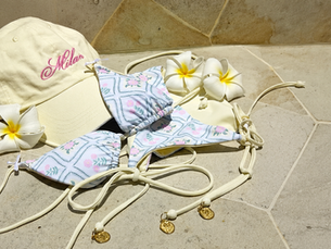 Beige cap with "Milan" text, floral bikini, and white plumeria flowers on light stone surface. Beachy and relaxed vibe.
