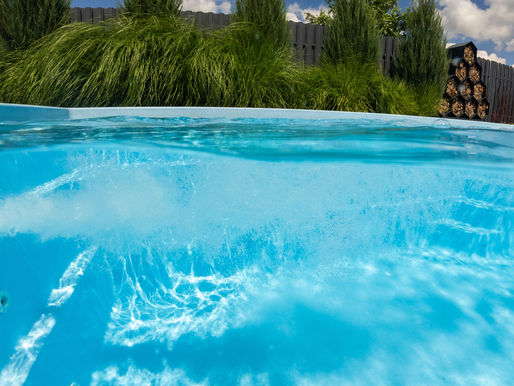 Blue hot tub water bubbling in a backyard, with tall grasses, a dark fence, stacked firewood, and a cloudy sky