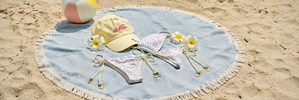 Beach scene with a blue towel, pastel bikini, yellow cap with "Milan" text, flowers, and a colorful beach ball on the sand. Relaxed mood.