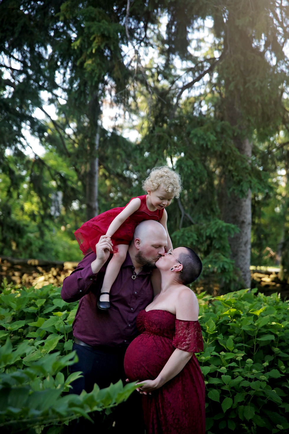 A man kisses a pregnant woman while holding a child on his shoulders in a forest. All wear red. The mood is joyful and loving.