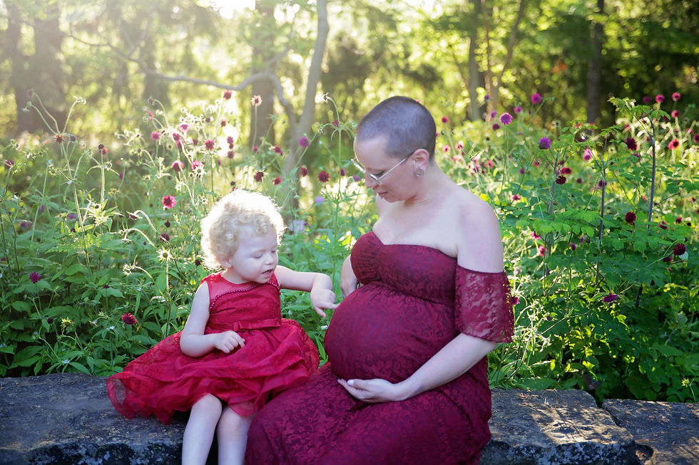 A child and a pregnant woman in red dresses sit on a stone ledge in a sunlit garden with purple flowers, sharing a tender moment.