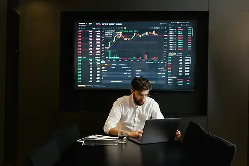 A man in a white shirt works on a laptop at a table, with financial charts and stock data displayed on a large screen behind him.