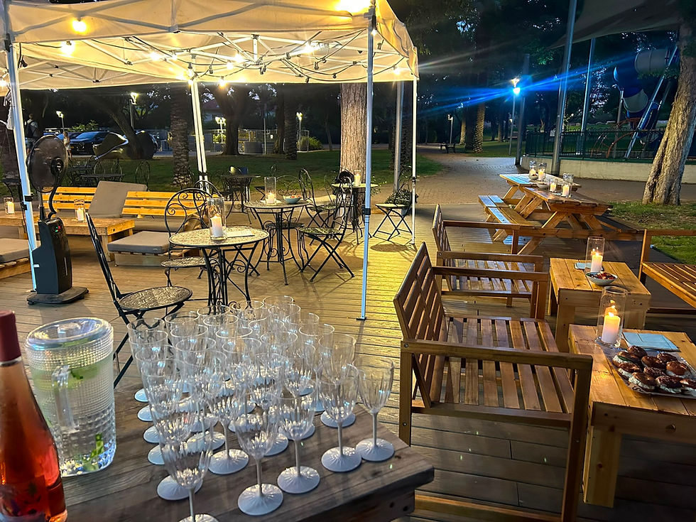 Evening outdoor event setup under a white canopy with string lights, wooden benches and tables, and many empty wine glasses on a wooden deck.