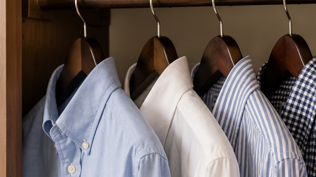 Classic men’s Oxford shirts in blue, white, pink and plaid hanging on wooden hangers in a warm, organized closet.