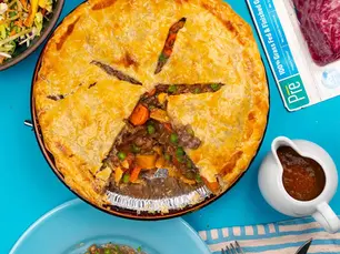 Top-down beef and vegetable pie on a blue table with salad and gravy; meat package reads 100% Grass-Fed & Finished Ground Beef