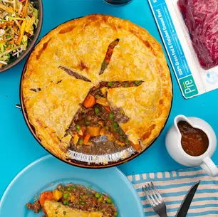 Top-down beef and vegetable pie on a blue table with salad and gravy; meat package reads 100% Grass-Fed & Finished Ground Beef