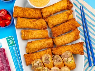 Plate of crispy egg rolls with dipping sauce, chopsticks, pickles, tomatoes, and a Pre ground beef pack on a bright blue table.