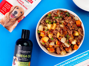 An overhead photo of a bowl of dog food, a water bowl, Pre 85% grass fed beef in the package and a package of Just Food For Dogs and Omega Oil.