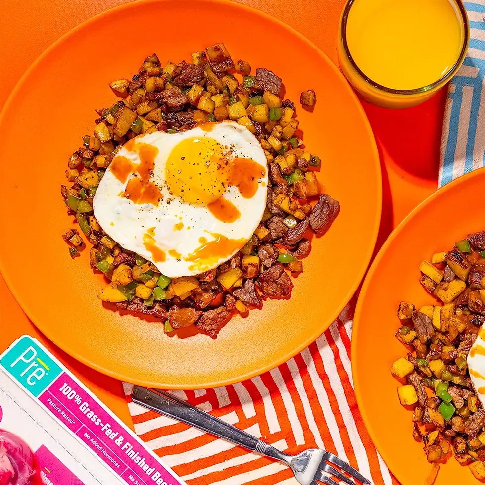 Orange plates of steak hash topped with a sunny-side-up egg, beside orange juice on a striped cloth and a Pre beef package.
