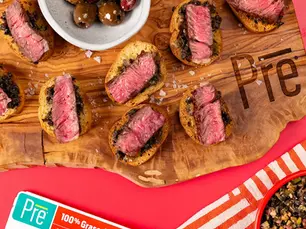 An overhead shot of steak slices on crostini bread and a bowl of olive tapenade nearby a package of Pre ribeye steak