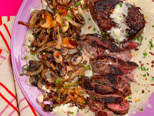 Sliced steak with sautéed mushrooms and creamy sauce on a purple plate, garnished with chives on a bright pink table.