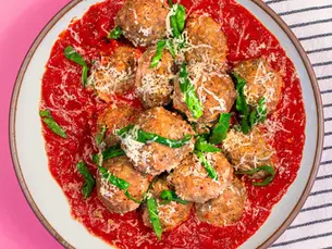 Overhead view of homemade grass-fed, pasture-raised beef meatballs in rich tomato sugo sauce, topped with grated Parmesan cheese and fresh basil on a white ceramic plate against a pink background.