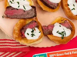 Beef-topped mini popovers with cream and herbs on a plate; Pre package below reads 100% Grass-Fed & Finish… on a red backdrop
