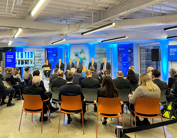 A professional panel discussion in a modern event space. Six speakers sit on stage, facing an attentive audience. Blue lighting and large screens decorate the room.