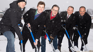 Five men in winter coats and suits participate in a snowy groundbreaking ceremony, smiling as they lean on shovels at a construction site.