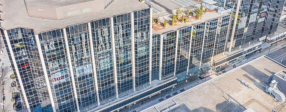 Aerial view of a modern building with a glass facade reflecting the cityscape. The rooftop features small garden areas, adding a touch of greenery.
