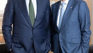 Deux hommes en costume bleu, debout et souriants dans un bureau avec de grandes fenêtres.