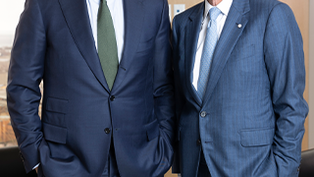 Deux hommes en costume bleu, debout et souriants dans un bureau avec de grandes fenêtres.