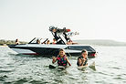 Two children in life vests sit on surfboards with an ATX boat behind them.