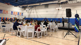 Banquet-style event in a gymnasium with speaker on stand addressing guests at round tables